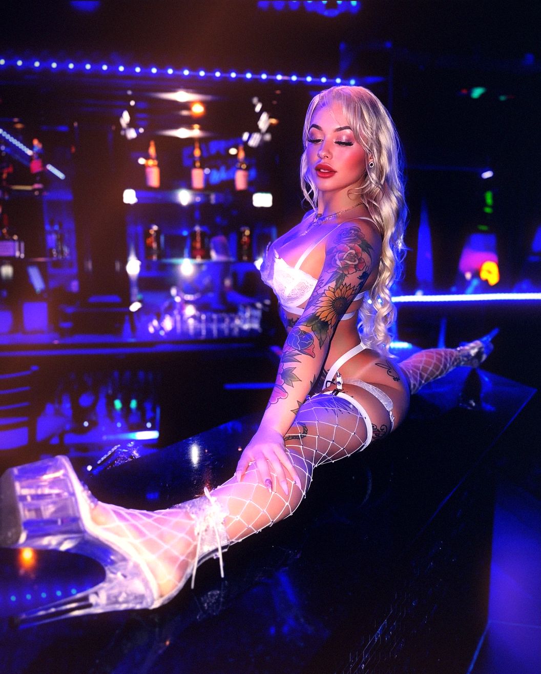 A performer sits along the polished black bar top in white lace and clear heels, the lit bottle wall stacked behind her and a blue LED rope running the length of the bar at Ten’s, the room dark beyond.