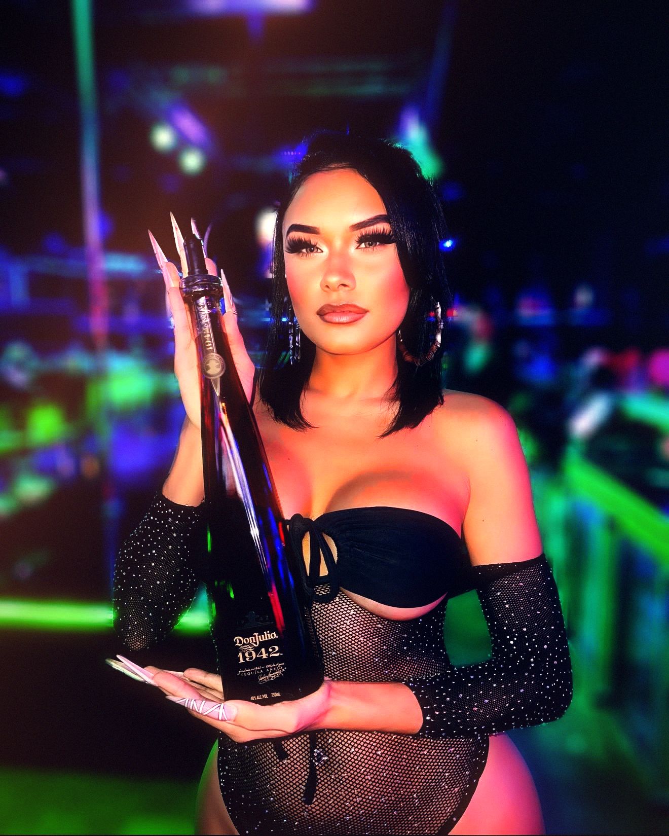 A host holds a tall dark bottle up beside her at the end of the bar, the room behind her going green and blue — the way bottle service arrives at a table at Ten’s.