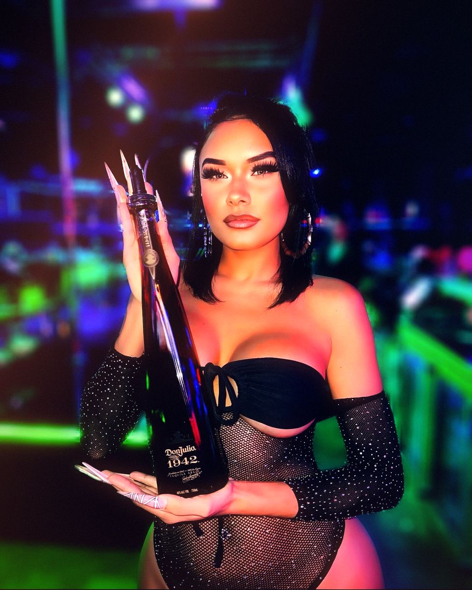 A host holds a tall dark bottle up beside her at the end of the bar, the room behind her going green and blue — the way bottle service arrives at a table at Ten’s.
