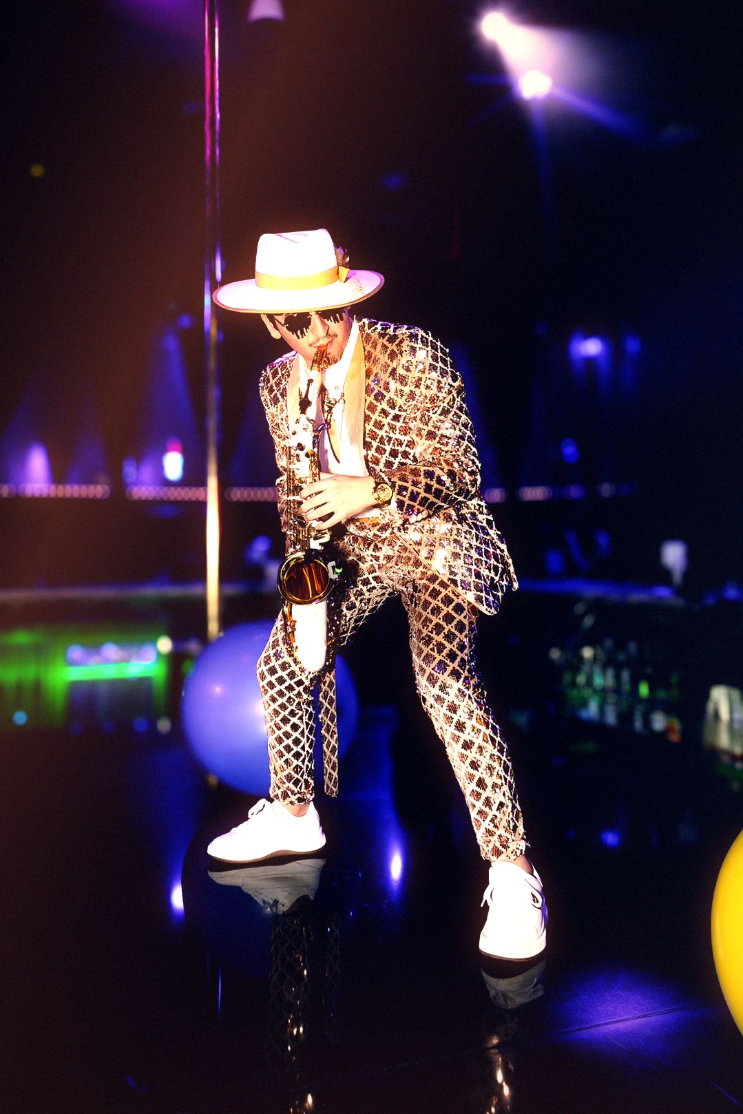 A saxophonist in a leopard-print sequinned suit, white brimmed hat and dark glasses plays beside the pole on Ten’s stage, the lit bottle wall behind him going green and violet under the spots.