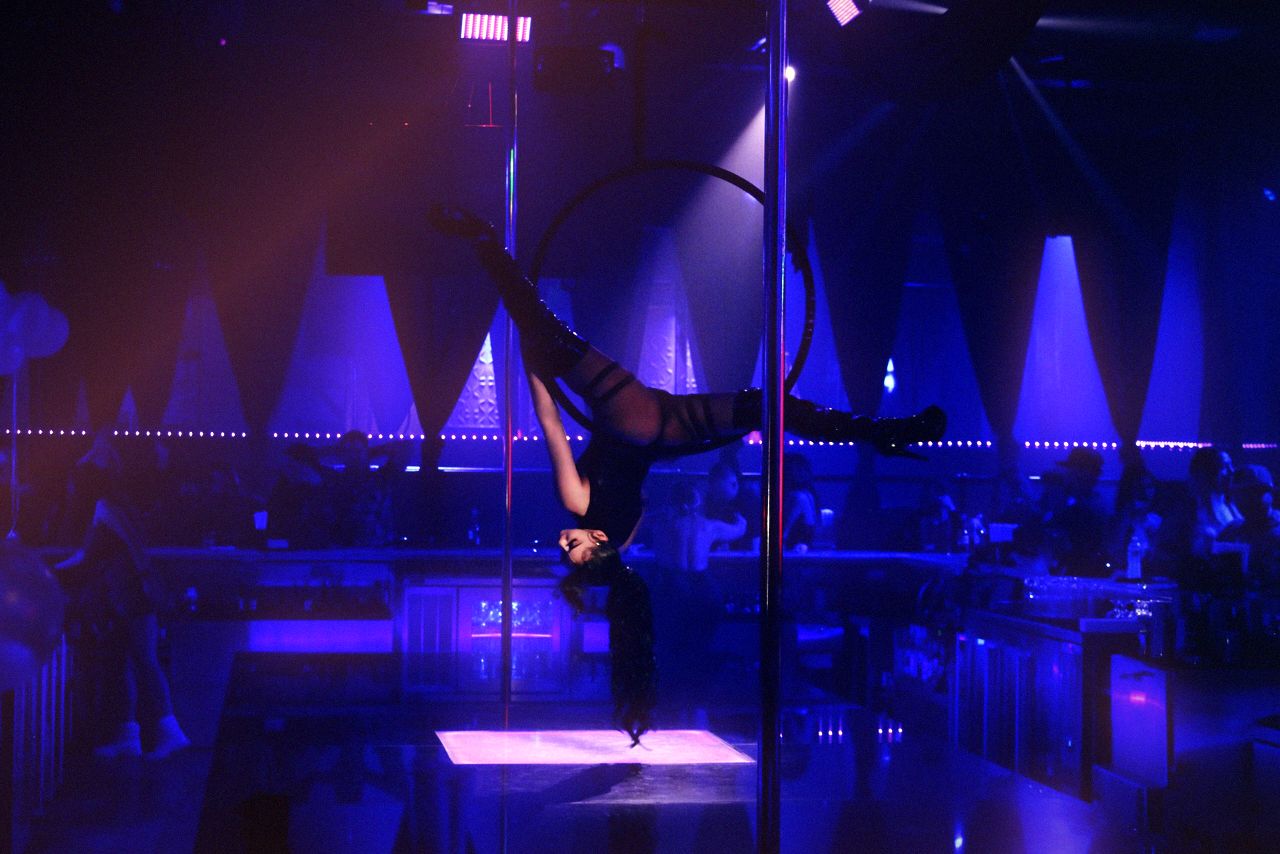 The main room at Ten’s seen from the floor: an aerialist inverted in a steel hoop rigged beside the chrome pole, a lit panel in the stage below her, white triangular fixtures along the back wall and a rope of pink lights running the length of the bar.