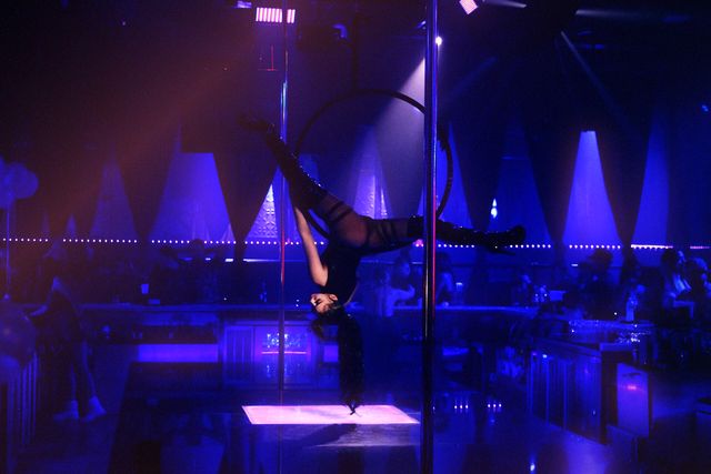 The main room at Ten’s seen from the floor: an aerialist inverted in a steel hoop rigged beside the chrome pole, a lit panel in the stage below her, white triangular fixtures along the back wall and a rope of pink lights running the length of the bar.